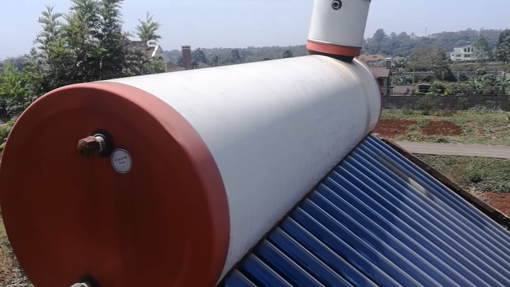 Close-up view of a rooftop passive solar water heater with a white storage tank, blue tubes, and a red end cap, set against a backdrop of urban landscape