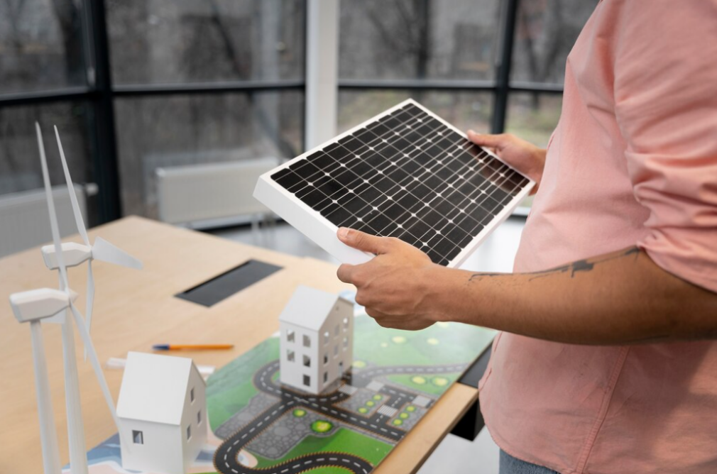 man in a pink shirt holds a solar panel in front of a table with small houses, a map, and two windmills  on the table