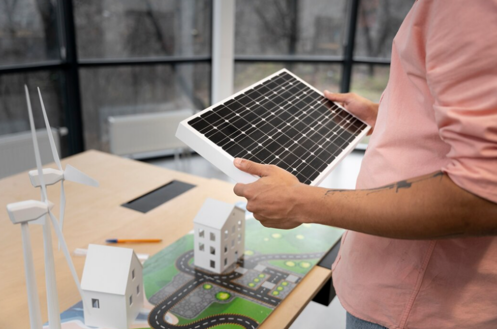 man in a pink shirt holds a solar panel in front of a table with small houses, a map, and two windmills on the table